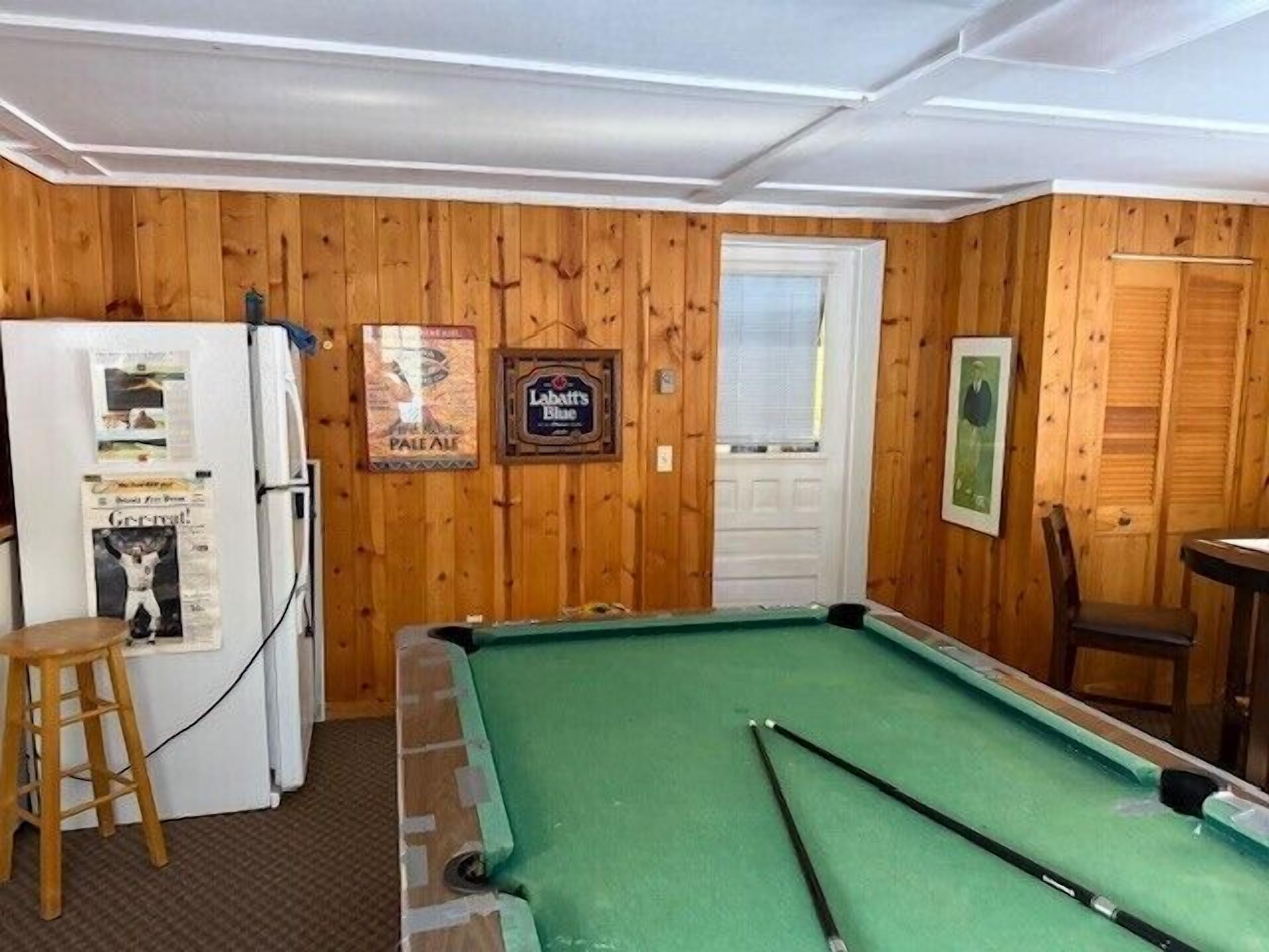 A pool table in a room with wooden walls, a refrigerator, and a door.