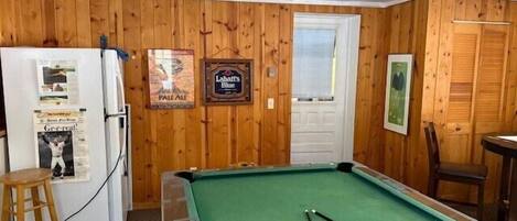 A pool table in a room with wooden walls, a refrigerator, and a door.
