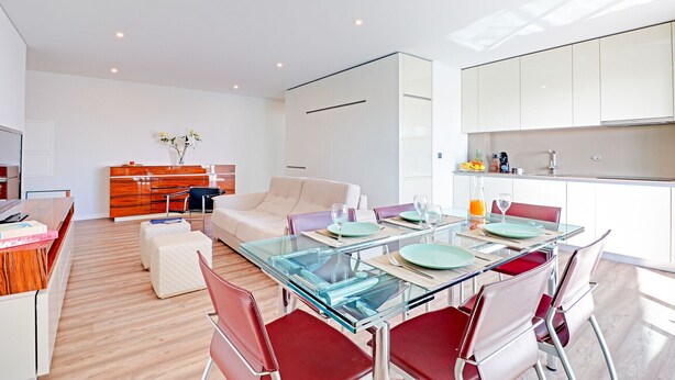 Una cocina moderna con armarios blancos, un comedor con mesas de vidrio y sillas rojas, y una sala de estar con un sofá y un mueble de madera.