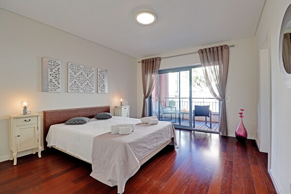 A bedroom with a large bed, a nightstand, a vase, and a view of the outside balcony.