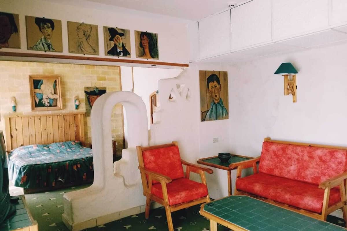 Una habitación pequeña y acogedora con una cama, dos sillones tapizados en rojo, una mesita y un tapete verde en el suelo.