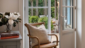 A wicker chair with a cushion, a vase with white flowers, and books on a table near a window. - The French House-Luxury Country Retreat (Trentham)
