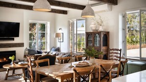 A cozy living room with a fireplace, a dining area with wooden furniture, and large windows offering a view of the outdoors. - The French House-Luxury Country Retreat (Trentham)