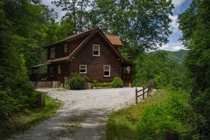 Un chalet rustique en bois, avec une allée en gravier, entouré d’une forêt dense et de collines.