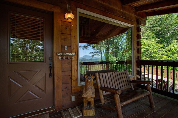 Une cabane en bois avec une pancarte « BIENVENUE », un banc et une vue sur les montagnes, visible à travers une grande fenêtre.