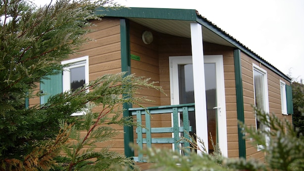 Una pequeña cabaña de madera con puerta y contraventanas verdes, rodeada de árboles.