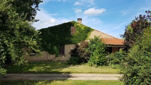 Uma casa rústica com telhado inclinado, rodeada por vegetação exuberante e um caminho de cascalho.