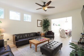 Un salon avec un canapé d’angle, une table basse sur laquelle est posé un jeu d’échecs, un ventilateur de plafond et, au fond, un coin repas.