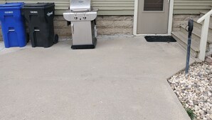 A concrete patio area with two trash bins, a barbecue grill, and a door. - Basement Studio Apartment (Sanborn)
