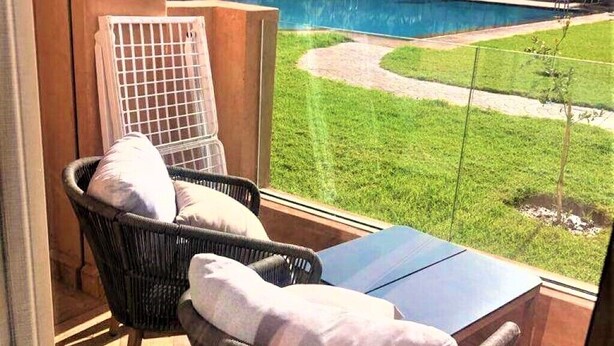 A balcony with a wicker chair and a table, overlooking a swimming pool and a grassy area. - Luxurious modern apartment Coralia Marrakech for rent (Marrakech)