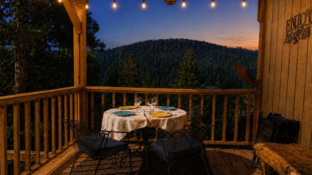 Une terrasse en bois avec une table dressée pour deux personnes, entourée de chaises, donnant sur une colline boisée.