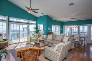 A spacious living room with a teal accent wall, large windows, a ceiling fan, and a dining area with white chairs and a wooden table.