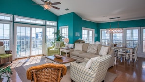 A spacious living room with a teal accent wall, large windows, a ceiling fan, and a dining area with white chairs and a wooden table. - Sea Grass Neighborhood * Private Pool 5 Bdrms (Cape San Blas)