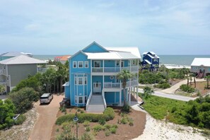 A two-story beachfront house with a balcony, a driveway, and a sandy beach in the background.