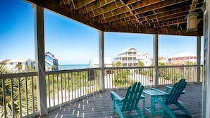 A covered outdoor area with a wooden deck, two green Adirondack chairs, and a view of the beach and ocean. - Sea Grass Neighborhood * Private Pool 5 Bdrms (Cape San Blas)
