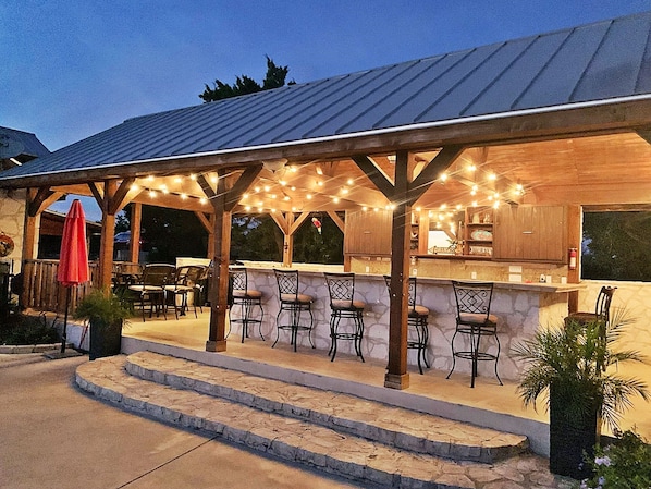 Uma área de bar ao ar livre com bancadas de pedra e banquetas, sob uma pérgola de madeira com luzes de cordão.