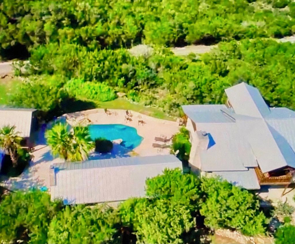 An aerial view of a property with a swimming pool surrounded by greenery, a covered patio area, and a large building with a sloped roof.