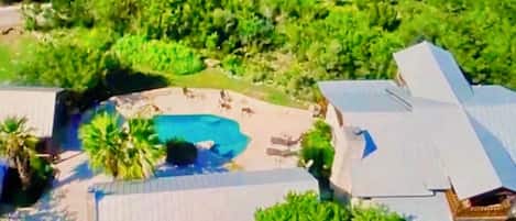Vista aérea de una propiedad con piscina rodeada de vegetación, un patio cubierto y un edificio grande con techo inclinado.