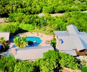 An aerial view of a property with a swimming pool surrounded by greenery, a covered patio area, and a large building with a sloped roof.