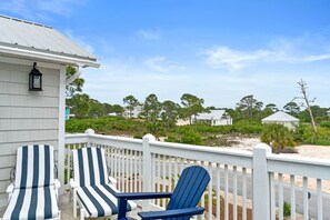 A deck with two striped chairs and a blue chair, overlooking a beach with houses and trees.