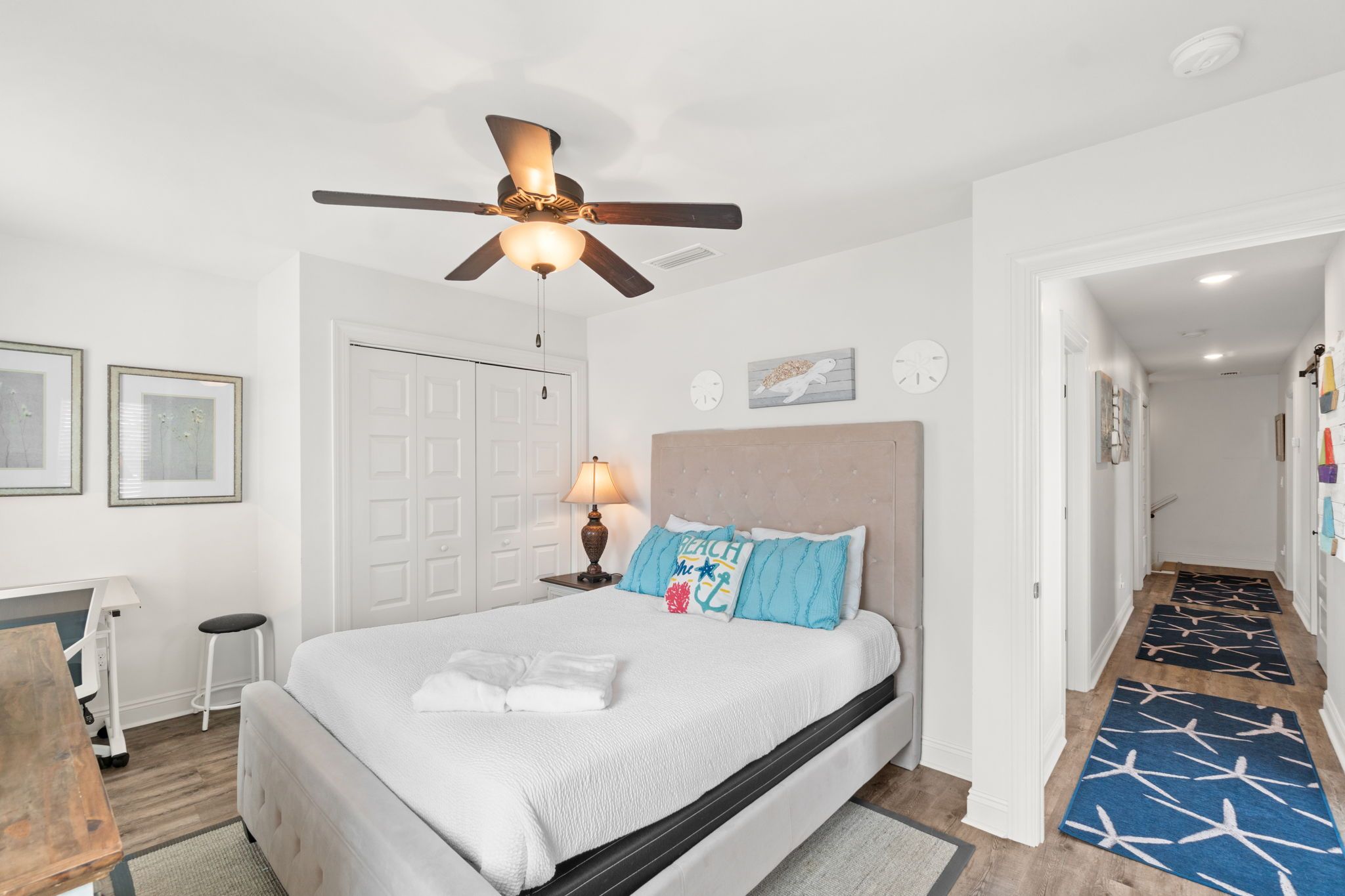 A bedroom with a bed, a ceiling fan, a bedside lamp, a door, and a hallway with a rug.