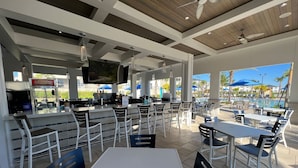 An outdoor dining area with white tables and chairs, a bar area with a TV, and a pool in the background. - Cinema Paradise: Last Minute Deal Private Pool/Spa (Davenport)