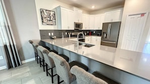A modern kitchen with white cabinets, a stainless steel refrigerator, a microwave, and a large island with bar stools. - Cinema Paradise: Last Minute Deal Private Pool/Spa (Davenport)