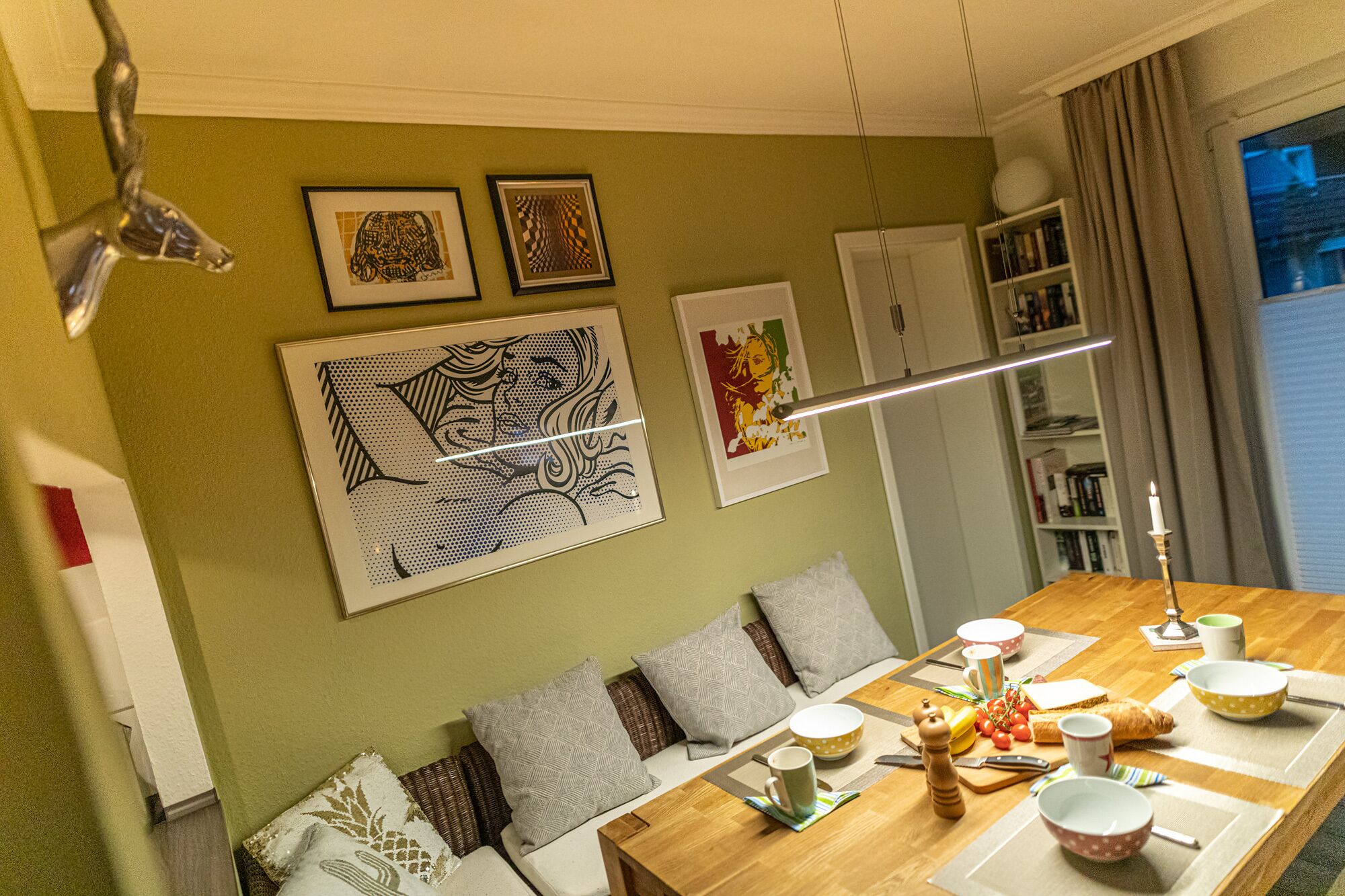 A dining area with a wooden table set for four, surrounded by cushioned seating. There are framed artworks on the wall, a bookshelf, and a window with curtains.