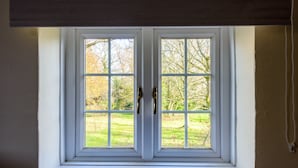 Ein Fenster mit weißen Rahmen und zwei Messinggriffen, das auf eine begrünte Fläche mit Bäumen blickt.