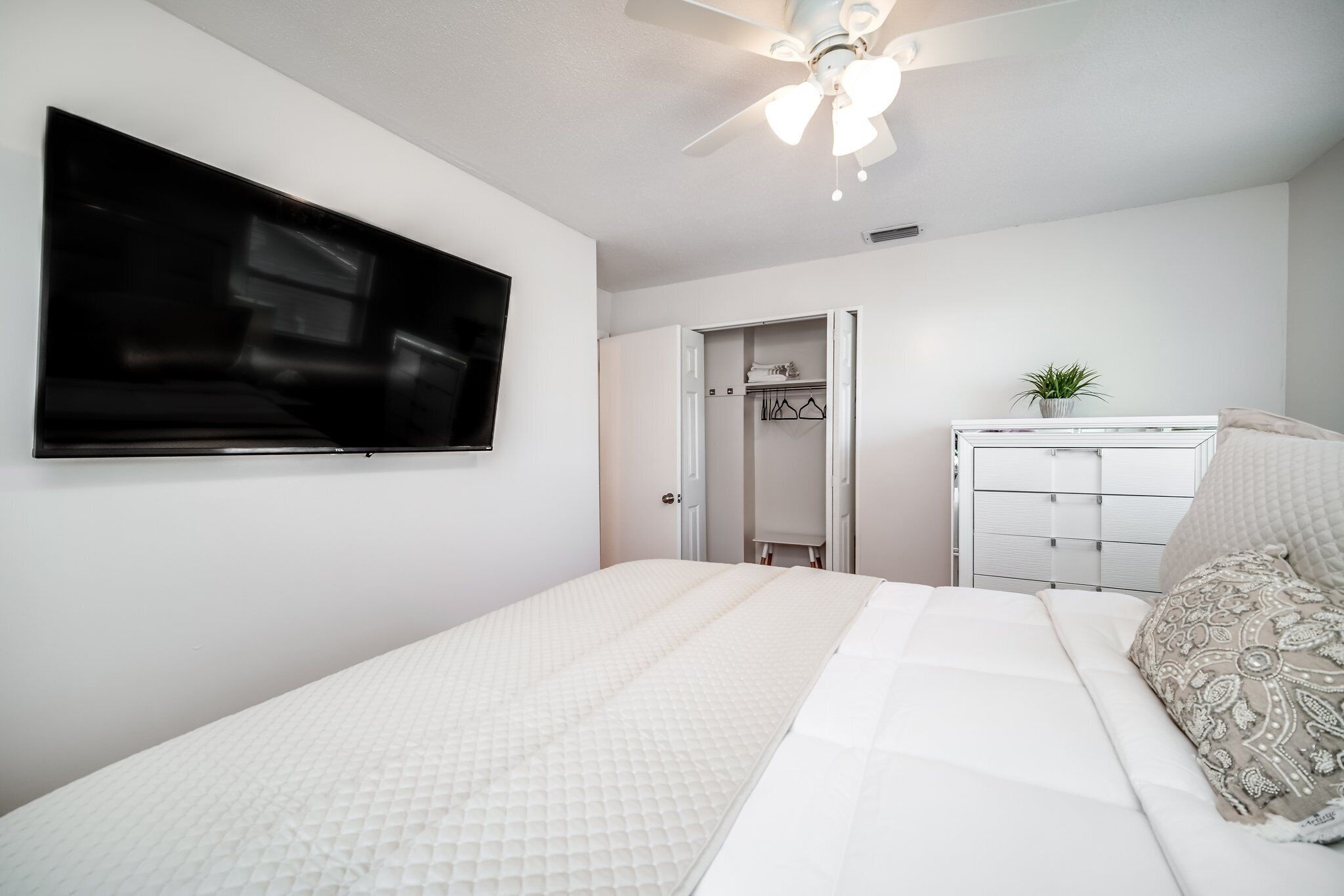 A bedroom with a bed, a TV mounted on the wall, a wardrobe, and a ceiling fan.