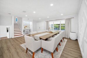 Un comedor amplio con una larga mesa de madera y sillas blancas tapizadas, una escalera que conduce a un nivel superior y vistas a follaje a través de amplios ventanales.