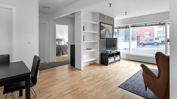 Un salon moderne avec une table à manger noire, un fauteuil marron et un téléviseur posé sur un meuble noir. Une grande fenêtre offre une vue sur la rue.