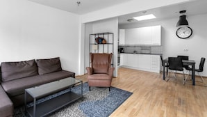 Un salon moderne avec un canapé en cuir marron, une table basse en verre et un tapis à motifs. On aperçoit un fauteuil marron et un coin repas avec une table et des chaises noires. La cuisine se trouve au fond, avec des meubles blancs et une horloge noire au mur.
