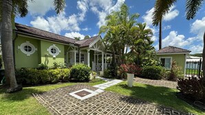 Ein grünes Haus mit einer Ziegeltterrasse und einem Garten mit Palmen und Sträuchern.