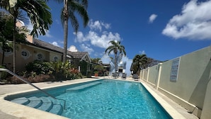 En swimmingpool med klart blåt vand, omgivet af palmer og en bygning med hvid facade.