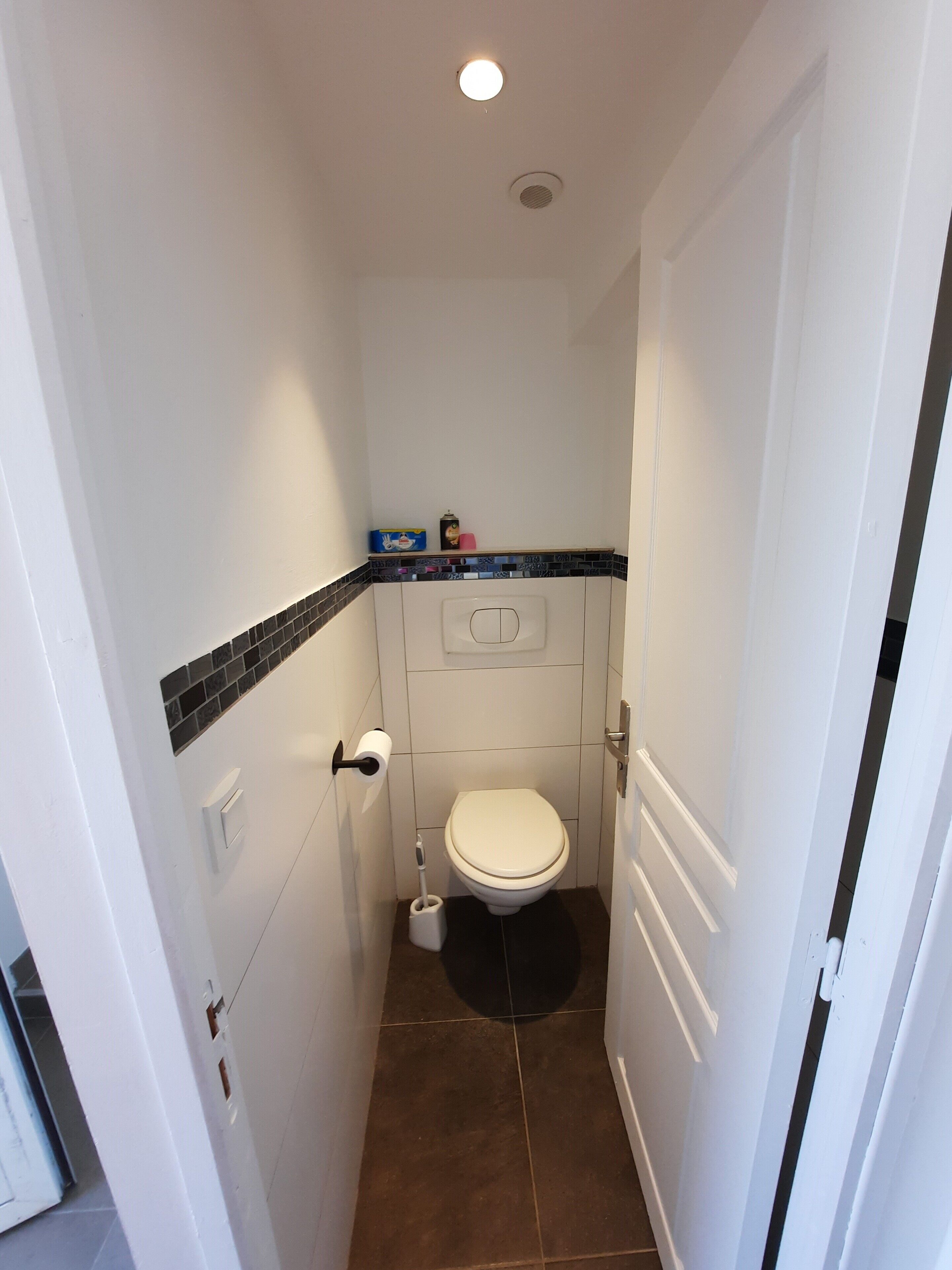 A narrow bathroom with a toilet, a small shelf with toiletries, and a tiled floor.