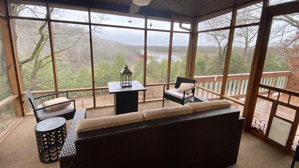 A covered patio with a sofa, chairs, and a table, overlooking a scenic view of trees and a body of water. - Large Home on Bull Shoals Lake with Core Lake Access (Midway)