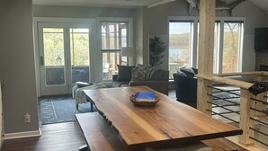 A spacious dining area with a large wooden table and benches, a cozy seating area with a sofa and chairs, and large windows offering views of the outdoors. - Large Home on Bull Shoals Lake with Core Lake Access (Midway)