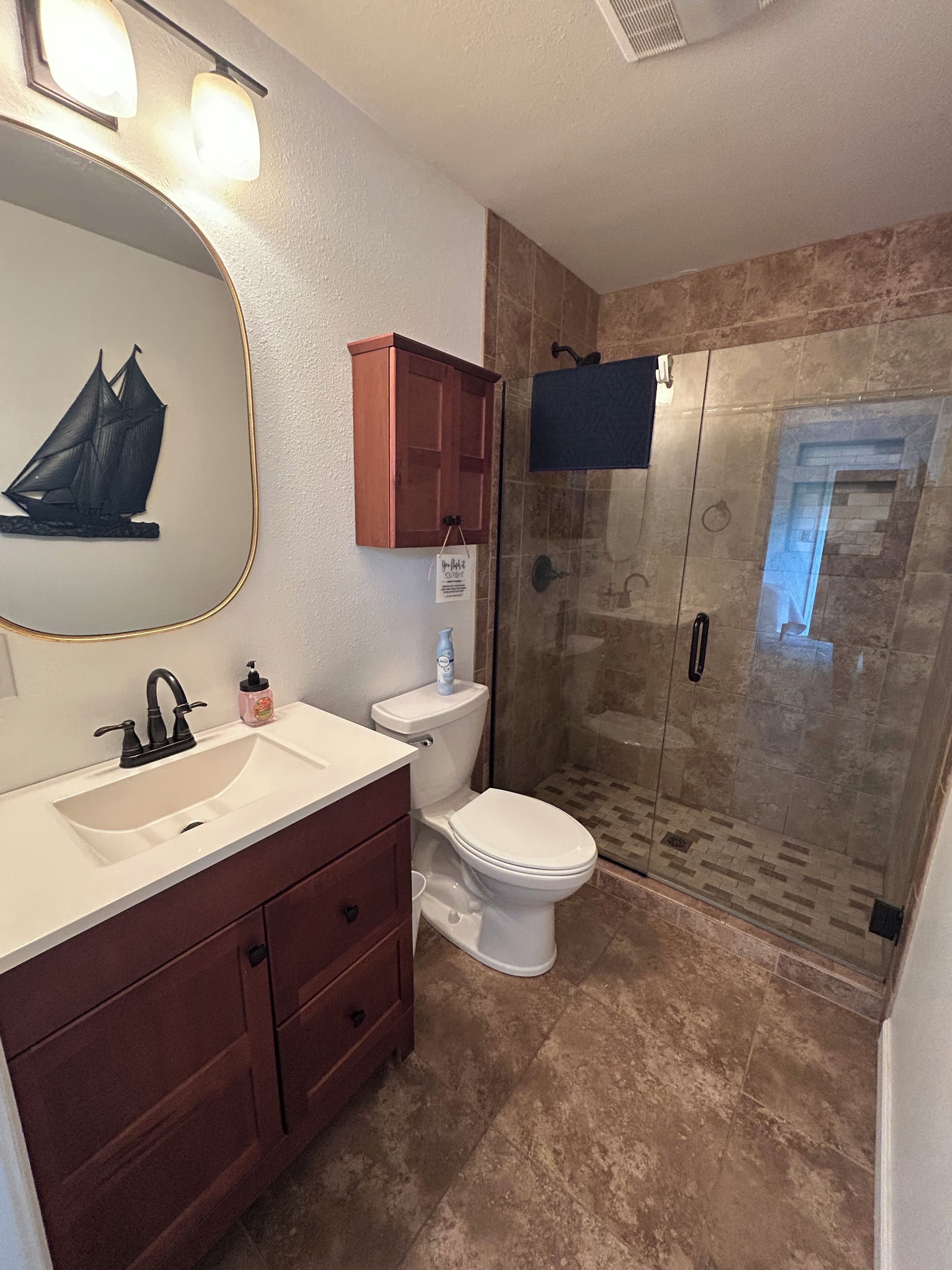 Une salle de bain avec un lavabo blanc, un robinet foncé, un meuble-lavabo en bois, des toilettes et une douche en verre avec un sol carrelé.