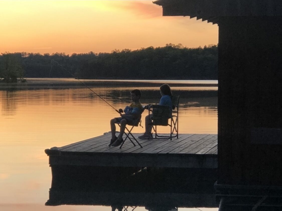 Two people sitting on chairs on a wooden dock by a calm lake during sunset.
