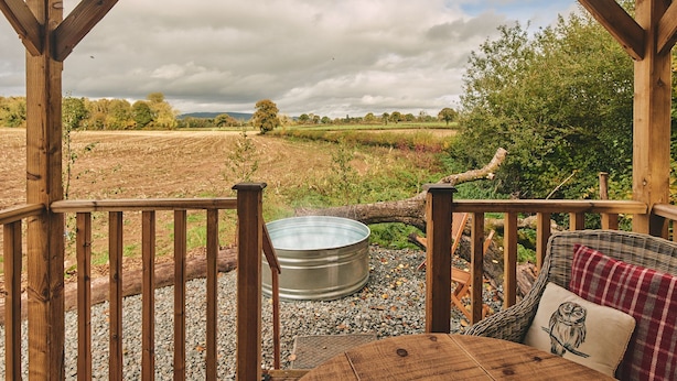 A wooden deck with a metal hot tub, wicker furniture, and a view of a field and trees.