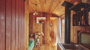 A wooden cabin interior with a wooden floor, a green sofa, a small table with flowers, a hanging light, and a kitchen area with a sink and stove.