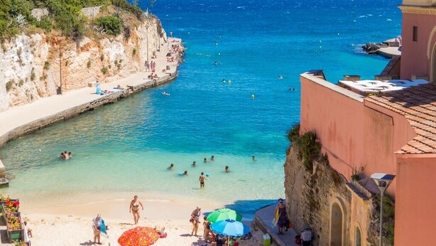 Una spiaggia costiera con acque blu cristalline, persone che nuotano e prendono il sole, e edifici con balconi che si affacciano sul mare.