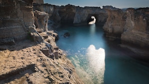 Paesaggio costiero con una formazione rocciosa naturale e una piccola struttura simile a una grotta.