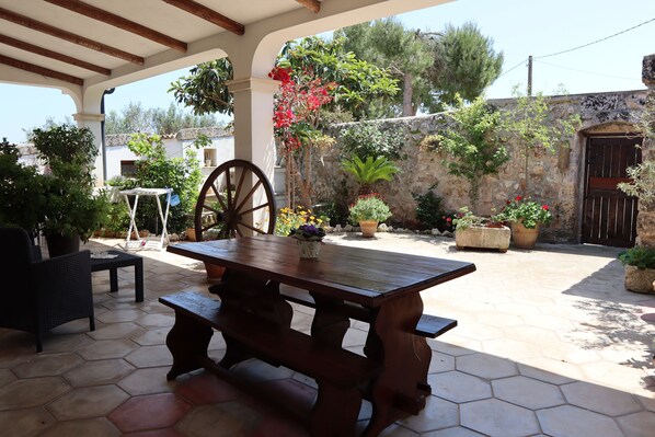 Una mesa de madera con bancos bajo una pérgola, rodeada de plantas en macetas y un muro de piedra.