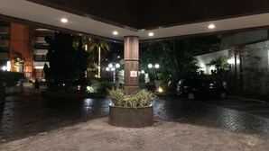 Um pátio interno com um vaso central, um carro preto estacionado à direita e um edifício com vários andares e varandas ao fundo.