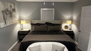 A bedroom with a bed, two bedside tables, a nightstand, a wall-mounted clock, a ceiling fan, and a door.