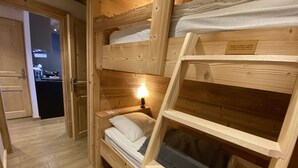 A wooden bunk bed room with a bedside table and lamp, a wooden door, and a kitchen area visible through an open door. - High standing, FOOT OF THE SLOPES and summer activities (Les Menuires)