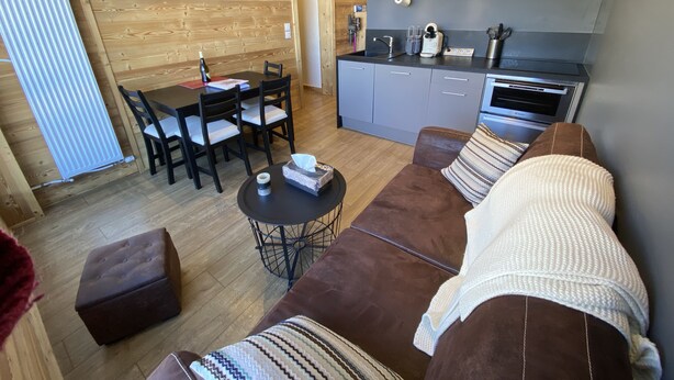 A cozy living room with a brown sofa, a small round table, and a kitchen area with grey cabinets and a built-in oven. - High standing, FOOT OF THE SLOPES and summer activities (Les Menuires)