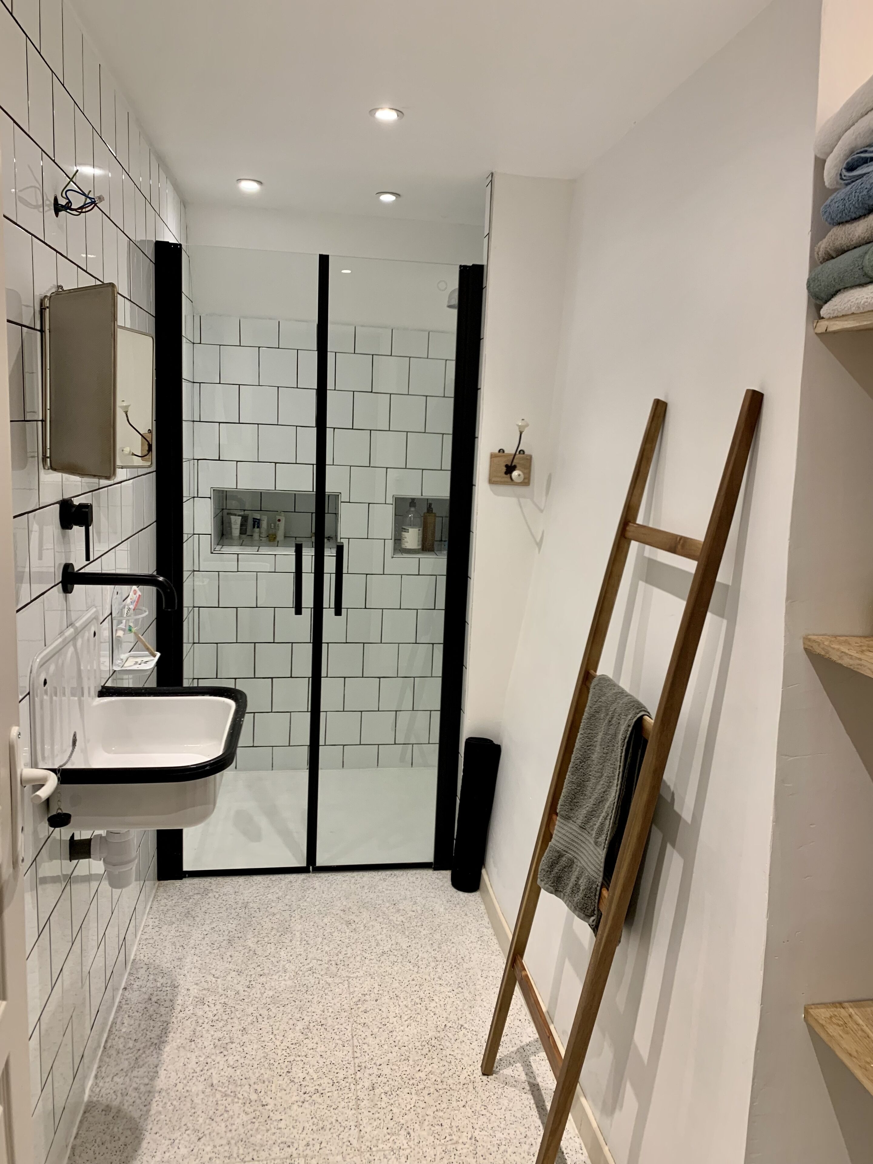 Een moderne badkamer met een glazen douchecabine, een witte wastafel met zwarte accenten en een houten ladder die tegen de muur staat.